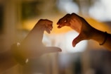 Two hands form a heart shape against a softly blurred background, bathed in warm, golden light. The image has a dreamy and intimate atmosphere, emphasizing connection and affection.