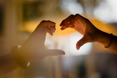 Two hands form a heart shape against a softly blurred background, bathed in warm, golden light. The image has a dreamy and intimate atmosphere, emphasizing connection and affection.