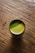 Close-up of a steaming cup of vibrant green matcha with delicate tea leaves beside it on a wooden table.