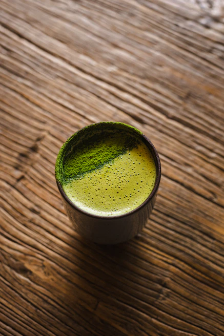 A steaming cup of green tea on a wooden table with fresh green tea leaves scattered around