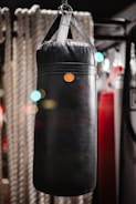 A black punching bag hangs prominently in a gym setting, surrounded by blurred ropes and gym equipment. Soft bokeh lights create a warm atmosphere in the background.