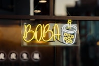 A neon sign featuring the word 'BOBA' in bright yellow letters, accompanied by a stylized image of a cup with bubbles, suggesting bubble tea. The sign is mounted on a window with darker background details.