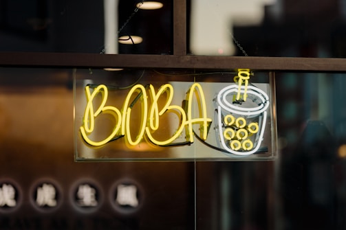 A neon sign featuring the word 'BOBA' in bright yellow letters, accompanied by a stylized image of a cup with bubbles, suggesting bubble tea. The sign is mounted on a window with darker background details.