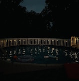 A dimly lit swimming pool surrounded by a white fence is visible at night. String lights are draped along the railing, casting a soft glow over the water. The scene includes several pool floaties, including a pumpkin-themed one, gently floating in the pool. Tall trees and a dark sky form the background, adding a serene and slightly mysterious atmosphere.