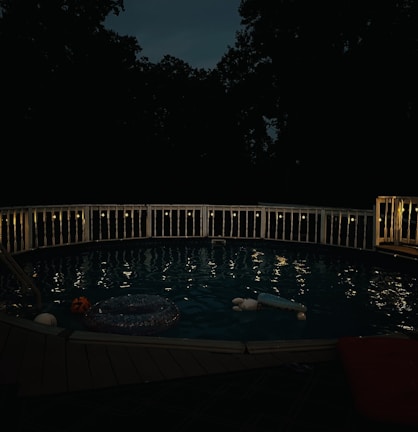 A dimly lit swimming pool surrounded by a white fence is visible at night. String lights are draped along the railing, casting a soft glow over the water. The scene includes several pool floaties, including a pumpkin-themed one, gently floating in the pool. Tall trees and a dark sky form the background, adding a serene and slightly mysterious atmosphere.
