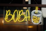 A bright neon sign featuring the word 'BOBA' in large yellow letters next to a neon depiction of a bubble tea cup filled with boba pearls, outlined in white and yellow. The sign is mounted behind a glass panel, with a dark and slightly reflective background.