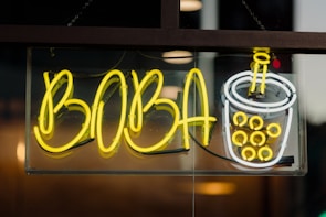 A bright neon sign featuring the word 'BOBA' in large yellow letters next to a neon depiction of a bubble tea cup filled with boba pearls, outlined in white and yellow. The sign is mounted behind a glass panel, with a dark and slightly reflective background.