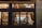 A window view into a cozy, modern restaurant featuring minimalistic interior design, warm lighting, and a neon sign displaying the name 'blossom.' The interior includes wooden shelves stocked with bottles, a row of tables and chairs, and an illuminated exit sign near a staircase.