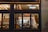 A window view into a cozy, modern restaurant featuring minimalistic interior design, warm lighting, and a neon sign displaying the name 'blossom.' The interior includes wooden shelves stocked with bottles, a row of tables and chairs, and an illuminated exit sign near a staircase.