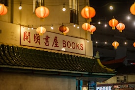 Red lanterns hang in rows, illuminating a scene at night. Below the lanterns, a building features a sign with both Chinese characters and the word 'BOOKS' in large red letters. The roof of the building has traditional Asian architectural elements, including green tiles. The surroundings suggest an urban area with a lively atmosphere.