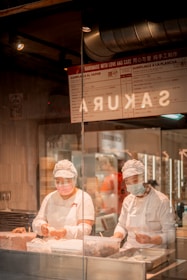 Two chefs wearing white uniforms and face masks are diligently preparing dumplings in a restaurant kitchen visible through a glass partition. The kitchen has a warm, inviting atmosphere with menu signage above, indicating offerings like dumplings al vapor. Industrial lighting and ventilation systems can be seen above the workspace.