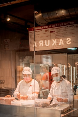 Two chefs wearing white uniforms and face masks are diligently preparing dumplings in a restaurant kitchen visible through a glass partition. The kitchen has a warm, inviting atmosphere with menu signage above, indicating offerings like dumplings al vapor. Industrial lighting and ventilation systems can be seen above the workspace.