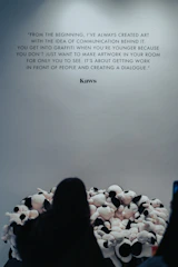 A wall features a prominent quote by Kaws about the importance of communication in art. In front of the wall, a person faces a large pile of plush toys or stuffed animals, creating an interactive art installation.