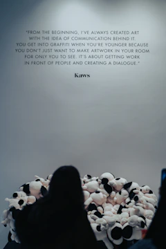 A wall features a prominent quote by Kaws about the importance of communication in art. In front of the wall, a person faces a large pile of plush toys or stuffed animals, creating an interactive art installation.