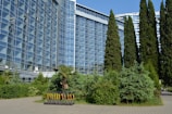 A modern glass building with reflective windows is surrounded by lush green trees and bushes. A row of electric scooters is neatly lined up on the sidewalk, contrasting with the natural greenery. Tall, slender trees tower in the background, adding to the urban landscape.