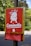 A metal box attached to a post features a red sign with a white cartoon illustration of a dog and the word 'Caninette' written underneath. Beneath the sign, a holder dispenses red plastic bags. The background is blurred, showing trees and pavement.