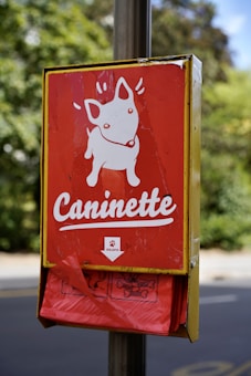 A metal box attached to a post features a red sign with a white cartoon illustration of a dog and the word 'Caninette' written underneath. Beneath the sign, a holder dispenses red plastic bags. The background is blurred, showing trees and pavement.