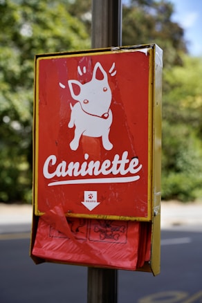 A metal box attached to a post features a red sign with a white cartoon illustration of a dog and the word 'Caninette' written underneath. Beneath the sign, a holder dispenses red plastic bags. The background is blurred, showing trees and pavement.