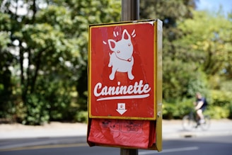 A bright red dog waste bag dispenser is mounted on a post against a backdrop of green foliage. The dispenser has a cartoon image of a dog above the word 'Caninette' and is partially encased in a yellow frame. A cyclist can be seen in the background on a path.