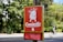 A bright red dog waste bag dispenser is mounted on a post against a backdrop of green foliage. The dispenser has a cartoon image of a dog above the word 'Caninette' and is partially encased in a yellow frame. A cyclist can be seen in the background on a path.