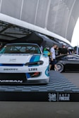 A custom-modified car with a white and blue paint job is displayed on a platform with a checkered design featuring branding logos such as LBWK and Nissan. In the background, there's a sleek black car partially visible, and several people are gathered near a tent-like structure. The setting appears to be an outdoor car show or exhibition with large architectural features overhead.