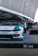 A custom-modified car with a white and blue paint job is displayed on a platform with a checkered design featuring branding logos such as LBWK and Nissan. In the background, there's a sleek black car partially visible, and several people are gathered near a tent-like structure. The setting appears to be an outdoor car show or exhibition with large architectural features overhead.