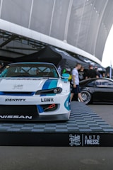 A custom-modified car with a white and blue paint job is displayed on a platform with a checkered design featuring branding logos such as LBWK and Nissan. In the background, there's a sleek black car partially visible, and several people are gathered near a tent-like structure. The setting appears to be an outdoor car show or exhibition with large architectural features overhead.