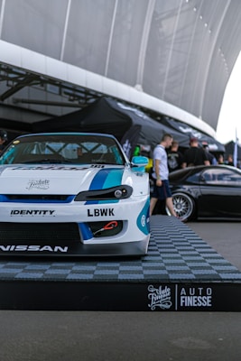 A custom-modified car with a white and blue paint job is displayed on a platform with a checkered design featuring branding logos such as LBWK and Nissan. In the background, there's a sleek black car partially visible, and several people are gathered near a tent-like structure. The setting appears to be an outdoor car show or exhibition with large architectural features overhead.