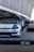 A custom-modified car with a white and blue paint job is displayed on a platform with a checkered design featuring branding logos such as LBWK and Nissan. In the background, there's a sleek black car partially visible, and several people are gathered near a tent-like structure. The setting appears to be an outdoor car show or exhibition with large architectural features overhead.