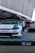 A custom-modified car with a white and blue paint job is displayed on a platform with a checkered design featuring branding logos such as LBWK and Nissan. In the background, there's a sleek black car partially visible, and several people are gathered near a tent-like structure. The setting appears to be an outdoor car show or exhibition with large architectural features overhead.