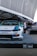 A custom-modified car with a white and blue paint job is displayed on a platform with a checkered design featuring branding logos such as LBWK and Nissan. In the background, there's a sleek black car partially visible, and several people are gathered near a tent-like structure. The setting appears to be an outdoor car show or exhibition with large architectural features overhead.