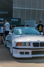 A white sports car with customized body modifications is parked in an urban setting. The car has a sleek, aerodynamic design with noticeable features including a front splitter and wide tires. Several people are casually standing nearby, some are focused on the vehicle, suggesting a car meet or exhibition. The background is urban with large structural elements.