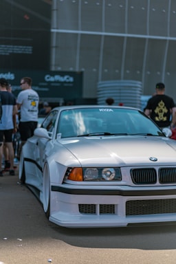 A white sports car with customized body modifications is parked in an urban setting. The car has a sleek, aerodynamic design with noticeable features including a front splitter and wide tires. Several people are casually standing nearby, some are focused on the vehicle, suggesting a car meet or exhibition. The background is urban with large structural elements.