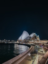 A night view of a famous opera house with its distinctive sail-shaped architecture illuminated against the night sky. The lights reflect off the water in the foreground. The scene includes a waterfront area with tables and seating, suggesting a nearby outdoor dining or bar area. In the background, city buildings are visible, adding to the urban setting.