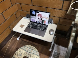 A compact study or workspace with a wooden panel backdrop features a small table holding a laptop displaying animated characters. There is a computer mouse beside the laptop, and a cushioned seat is positioned in front of the table. The setting suggests a cozy and personalized work environment, with a curtain partially visible on the right side.