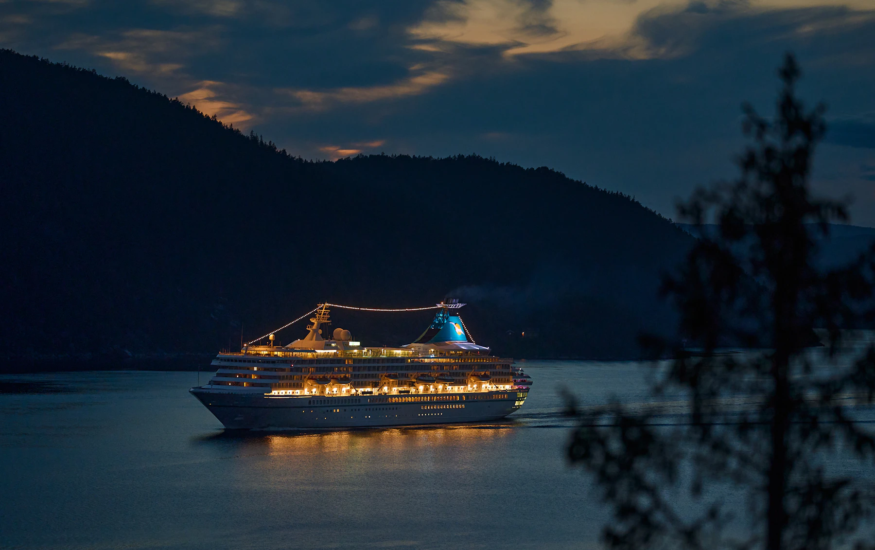 A small dark-hulled ship at anchor in a fjord at dusk, lights on, hills behind