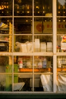 A view through a multi-paned window showing the interior of a shop or restaurant. The window is slightly dirty, and through it, stacks of plastic cups, white containers, and various cleaning supplies are visible on shelves and counters. The background includes a sign with partially visible text and a colorful flyer pinned to the window frame. Light bulbs and reflections can be seen through the glass.
