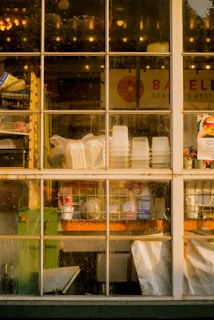 A view through a multi-paned window showing the interior of a shop or restaurant. The window is slightly dirty, and through it, stacks of plastic cups, white containers, and various cleaning supplies are visible on shelves and counters. The background includes a sign with partially visible text and a colorful flyer pinned to the window frame. Light bulbs and reflections can be seen through the glass.