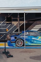 A blue sports car with detailed graphics is parked next to a trailer. The car has a large rear spoiler and a visible brand logo on the side. A hydraulic jack is placed nearby on the concrete pavement. The trailer has a dark color scheme with angular design elements.