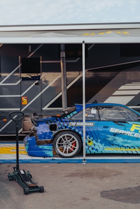 A blue sports car with detailed graphics is parked next to a trailer. The car has a large rear spoiler and a visible brand logo on the side. A hydraulic jack is placed nearby on the concrete pavement. The trailer has a dark color scheme with angular design elements.