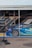 A blue sports car with detailed graphics is parked next to a trailer. The car has a large rear spoiler and a visible brand logo on the side. A hydraulic jack is placed nearby on the concrete pavement. The trailer has a dark color scheme with angular design elements.