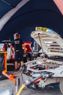A workshop scene where a white rally car is undergoing maintenance or repair. The car's hood is open, revealing intricate engine and mechanical parts. Several people are working around the vehicle, with tools and equipment scattered around. The atmosphere is busy, and the scene is set under a tent-like structure, likely in a pit area.