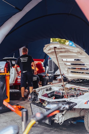 A workshop scene where a white rally car is undergoing maintenance or repair. The car's hood is open, revealing intricate engine and mechanical parts. Several people are working around the vehicle, with tools and equipment scattered around. The atmosphere is busy, and the scene is set under a tent-like structure, likely in a pit area.