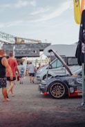 A group of people are gathered around a modified gray sports car with an open hood, displaying various brand stickers and decals. In the background, there are industrial structures and several other vehicles. The scene is outdoors, likely at an event or car show.