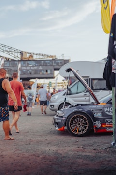 A group of people are gathered around a modified gray sports car with an open hood, displaying various brand stickers and decals. In the background, there are industrial structures and several other vehicles. The scene is outdoors, likely at an event or car show.