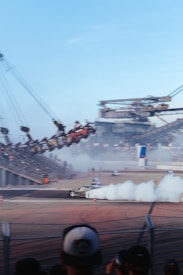 A car is drifting on a racetrack, producing a large plume of white smoke. Spectators are seated on bleachers in the background, observing the event. In the distance, industrial machinery and structures are visible, adding a dramatic backdrop.
