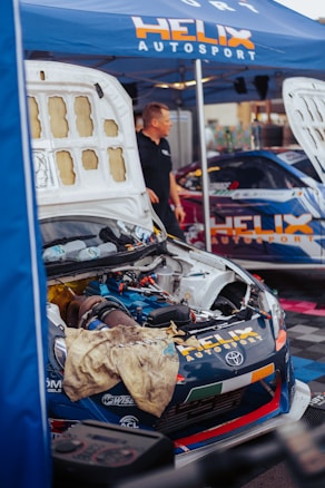 A car with its hood open is parked under a blue tent labeled 'HELIX AUTOSPORT'. The engine is exposed, with a dirty cloth draped over the front. A person stands nearby, wearing a black shirt. Another car is partially visible in the background, also branded with similar logos.