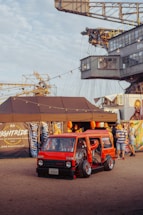 A custom red minivan with its doors open displays an elaborate interior setup. The scene is set in an outdoor festival environment with people gathered around. A large industrial crane looms in the background, and string lights provide a festive atmosphere. Various banners and a tent setup add to the event-like setting.