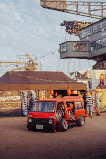 A custom red minivan with its doors open displays an elaborate interior setup. The scene is set in an outdoor festival environment with people gathered around. A large industrial crane looms in the background, and string lights provide a festive atmosphere. Various banners and a tent setup add to the event-like setting.