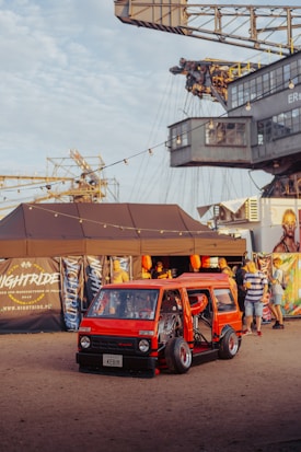 A custom red minivan with its doors open displays an elaborate interior setup. The scene is set in an outdoor festival environment with people gathered around. A large industrial crane looms in the background, and string lights provide a festive atmosphere. Various banners and a tent setup add to the event-like setting.